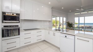 Una cocina moderna con armarios blancos, horno empotrado, microondas y lavavajillas. Tiene un ventanal con vistas a la playa.