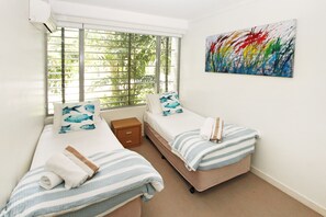 Un dormitorio con dos camas, una ventana con persianas, una mesita de noche pequeña y un cuadro abstracto y colorido en la pared.