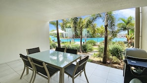 A dining area with a glass table and chairs, a grill, and a view of a pool and palm trees. - Blue on Blue Apartment 815 (Nelly Bay)