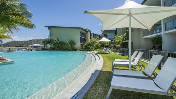 A swimming pool with lounge chairs and umbrellas, surrounded by a building with balconies and a view of the beach. - Blue on Blue Apartment 815 (Nelly Bay)