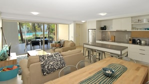 A modern living room with a sofa, coffee table, and dining table. There is a kitchen area with a refrigerator, sink, and cabinets. A sliding glass door leads to a balcony with a pool and palm trees. - Blue on Blue Apartment 815 (Nelly Bay)