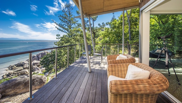 Eine Holzterrasse mit einem Korbsessel und Sesseln, mit Blick auf eine felsige Küstenlinie und das Meer.