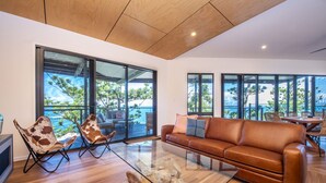A spacious living area with a leather sofa, a glass coffee table, and a wooden floor. Large windows offer views of the outdoors. - Headland House (Picnic Bay)