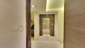 A hallway with an elevator, a staircase, and a wall-mounted light fixture. - Hotel Grandeur (Pune)