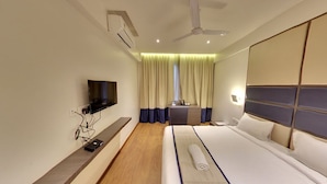A modern hotel room with a large bed, a flat-screen TV mounted on the wall, a ceiling fan, and a window with curtains. - Hotel Grandeur (Pune)