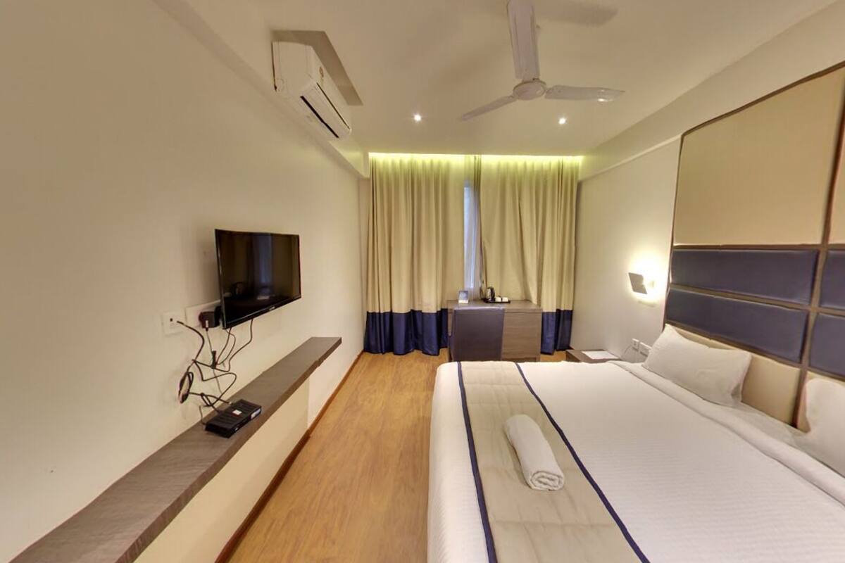A modern hotel room with a large bed, a flat-screen TV mounted on the wall, a ceiling fan, and a window with curtains.