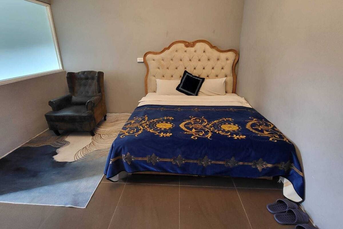 Un dormitori amb un capçal tapissat, un armchair fosc, un edredó estampat i un parell d'esmilles a terra.