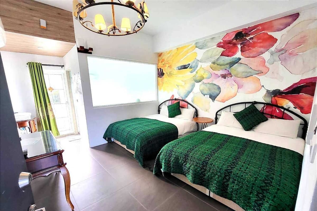 Habitació amb dos llits, un mural floral gran, un penjador de cristall i una finestra amb cortines.