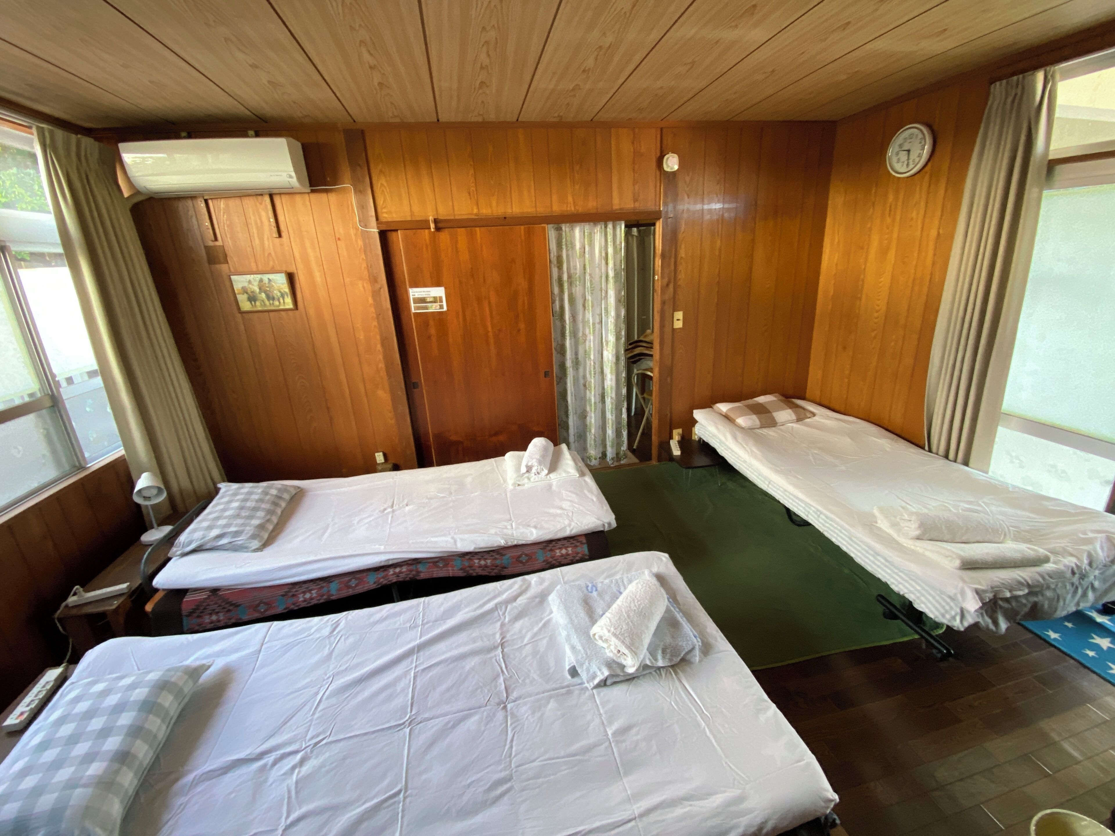 A room with two beds, wooden walls, a clock, and a window with curtains.