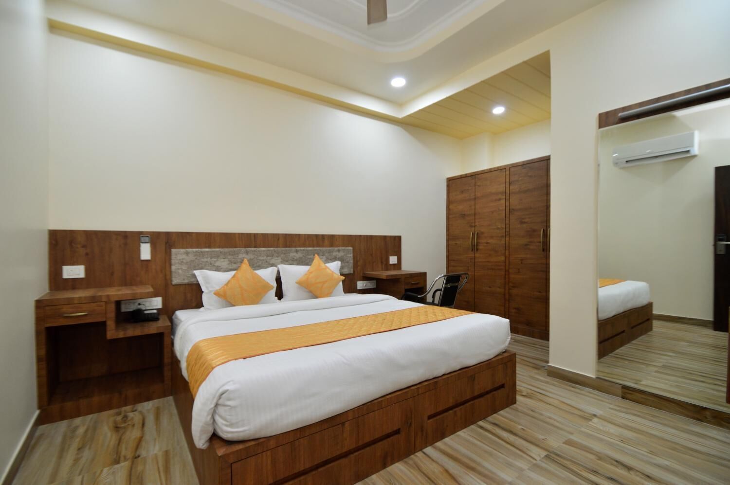 A hotel room with a large bed, wooden headboard, a desk with a chair, a wardrobe, and a mirror.