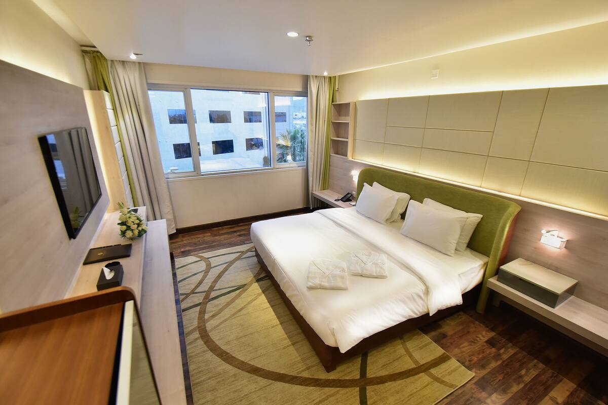 A hotel room with a bed, a desk, a TV, and a window with a view of buildings.