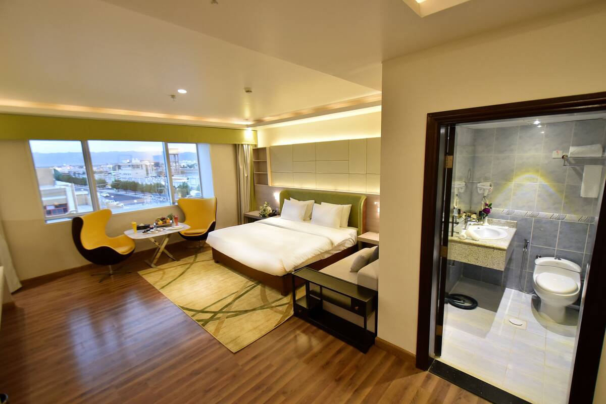 A modern hotel room with a large bed, a seating area with a chair, a small table, and a view of the city through large windows.
