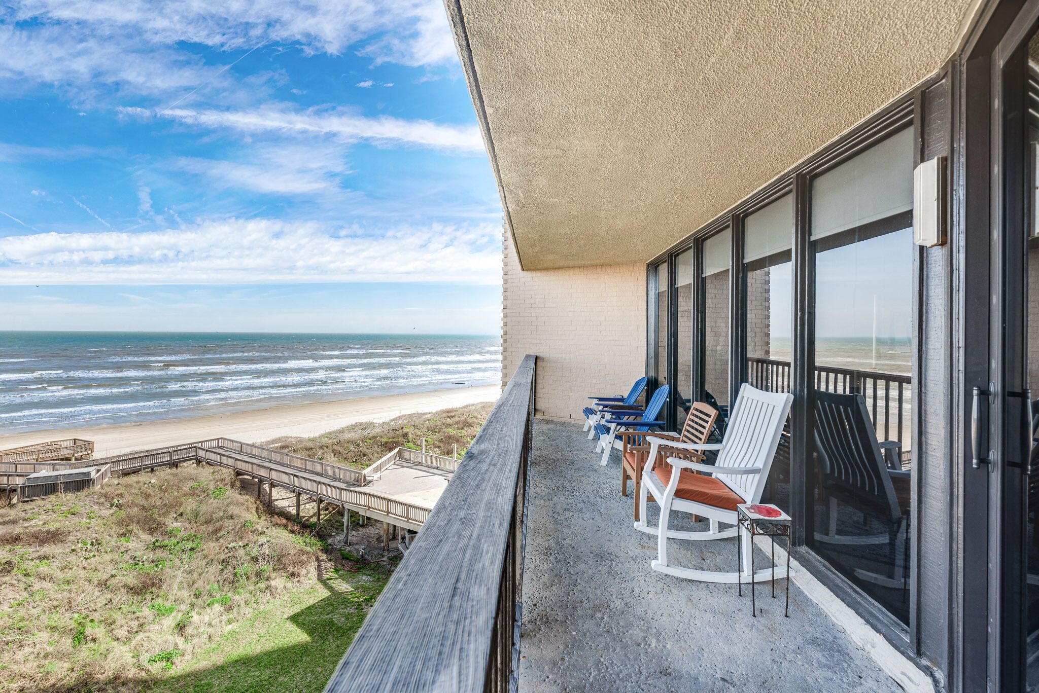 A balcony with a view of the beach and ocean, featuring several chairs and a wooden deck.