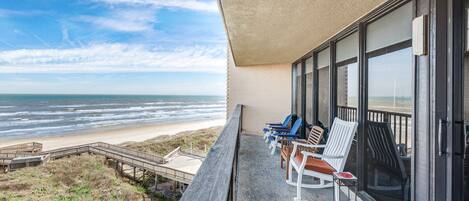A balcony with a view of the beach and ocean, featuring several chairs and a wooden deck.
