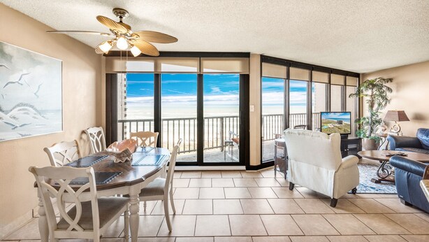 A spacious living area with a large window overlooking the beach, a dining table with chairs, a sofa, and a ceiling fan.