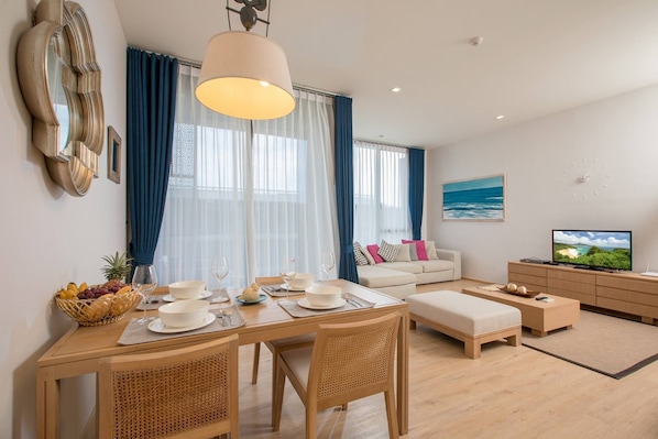 A modern living room with a dining area, a sofa set, a television, and a large window with curtains.