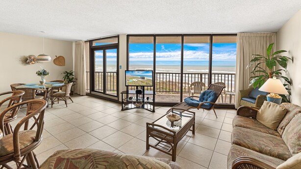 Ein geräumiges Wohnzimmer mit Balkon und Blick auf den Strand, ausgestattet mit Korbstühlen und einer Couch.