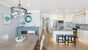 A modern kitchen with white cabinets, a central island, and a dining area with a wooden table and chairs. - Sea Gull #808: Beachfront 2 Bedroom 2 Bath with Spectacular Views and 24 Hour Management (Port Aransas)