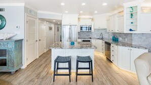 A modern kitchen with white cabinetry, a marble countertop, and stainless steel appliances. There are two bar stools and a white chair. - Sea Gull #808: Beachfront 2 Bedroom 2 Bath with Spectacular Views and 24 Hour Management (Port Aransas)