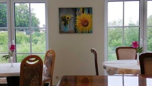 A dining area with a wooden table and chairs, a painting of sunflowers on the wall, and large windows with a view of greenery.