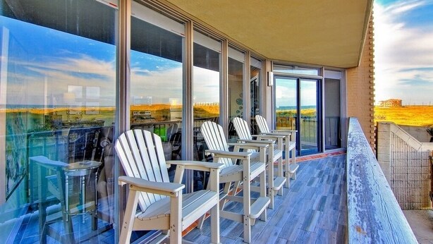 Un balcón con sillas Adirondack blancas y barandilla de vidrio que ofrece vistas a un paisaje pintoresco.