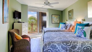 A bedroom with two beds, a ceiling fan, a television, and a balcony with a view of greenery.