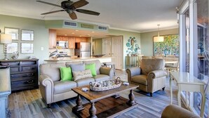 A spacious living room with a sofa, armchairs, and a coffee table. A ceiling fan and kitchen are visible in the background.