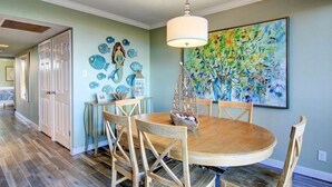 A dining area with a round wooden table, wooden chairs, a hanging light fixture, and a large abstract painting on the wall.