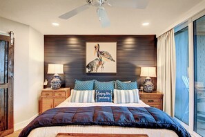 A bedroom with a large bed, a ceiling fan, a wall-mounted painting of a bird, and two bedside tables with lamps.