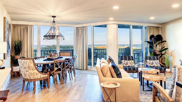 Amplia sala de estar con mesa de comedor, sofás y sillas. Grandes ventanales ofrecen vistas al exterior.