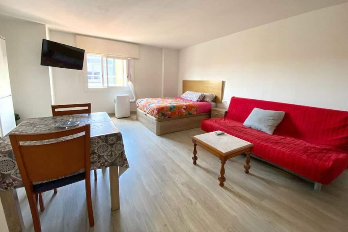 Quarto compacto com cama, sofá vermelho, mesa de jantar com cadeiras e TV de tela plana presa à parede.