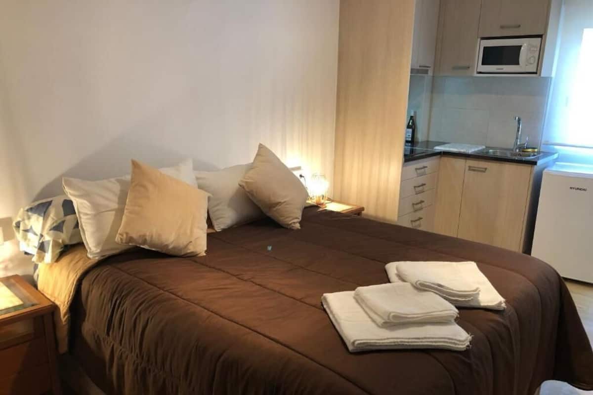 Cama bem arrumada com roupa de cama marrom, mesa de cabeceira com luminária e cozinha com micro-ondas e pia.