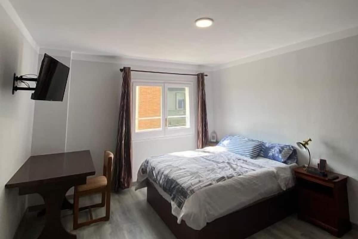 Quarto com cama, escrivaninha, cadeira, televisão, janela com cortinas e mesa de cabeceira com abajur.