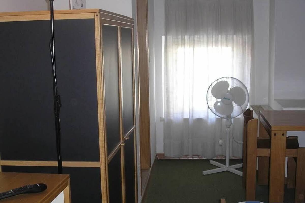 Een kleine kamer met een bed, een bureau, een stoel, een ventilator en een scheidingswand.