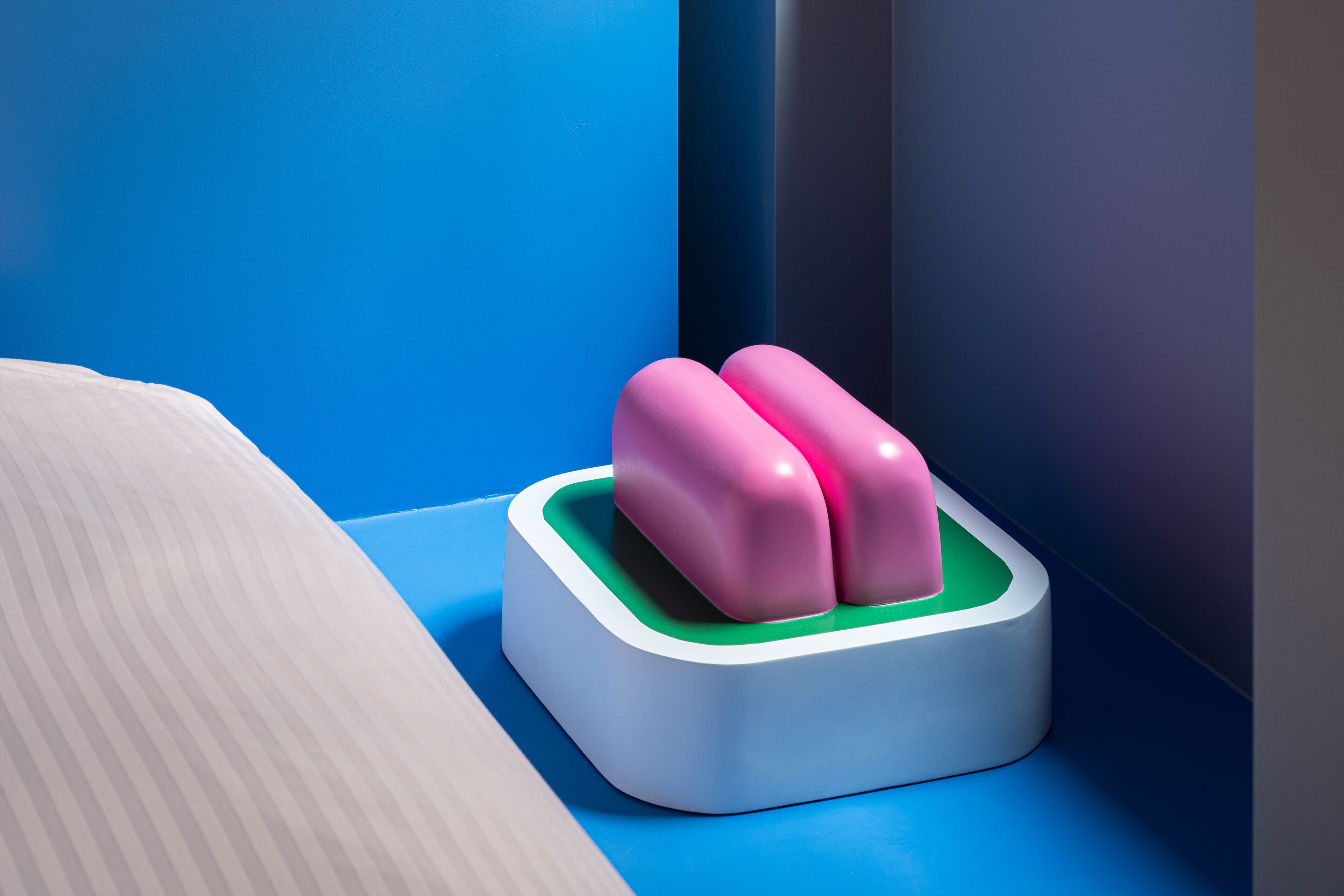 Un lit avec une tête de lit blanche et un oreiller, une table de chevet blanche, et un objet rose et vert sur le sol.