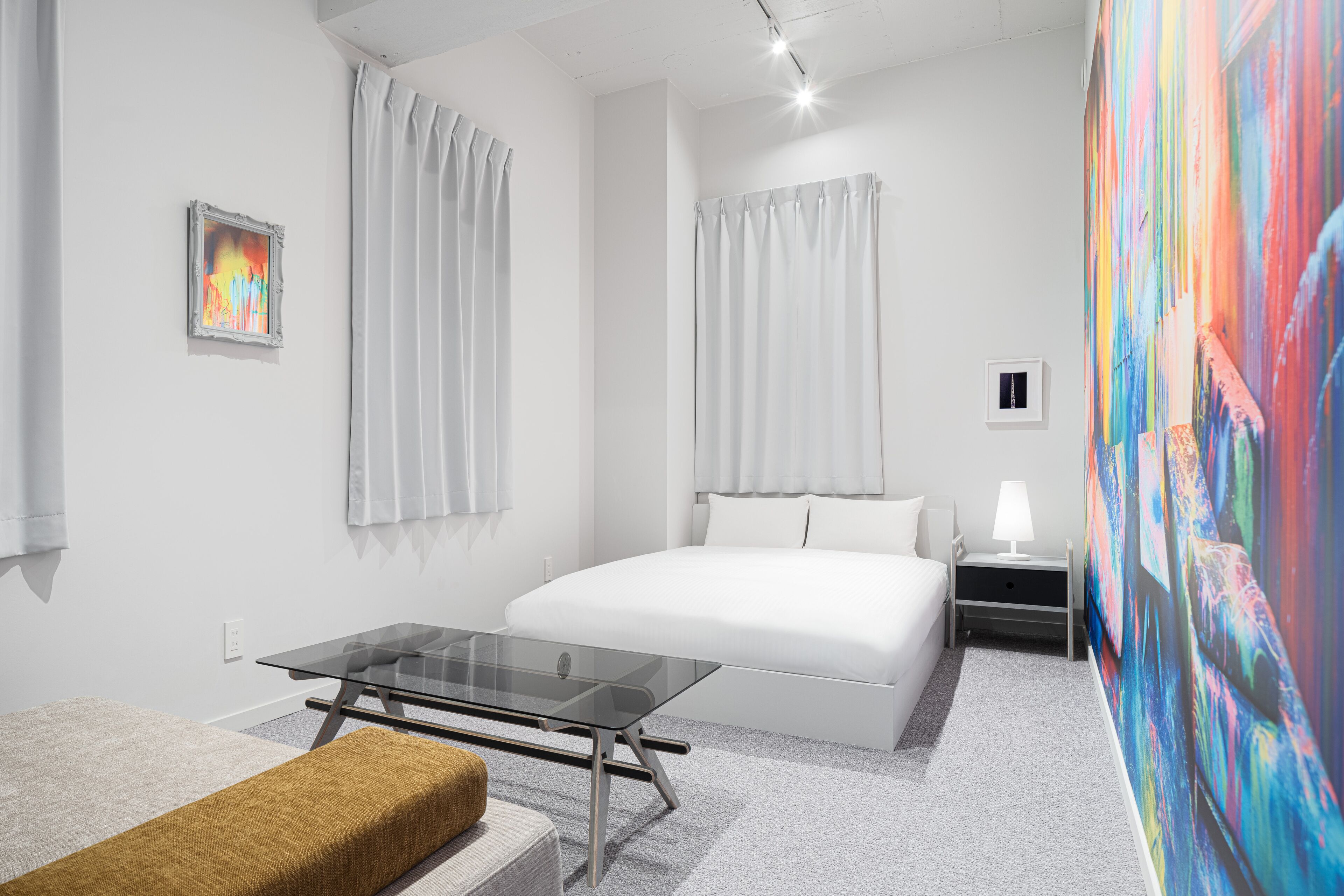 A modern hotel room with a large bed, a glass table, a bench, and a colorful abstract painting on the wall.