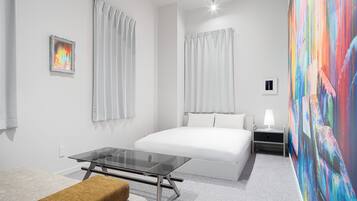 A modern hotel room with a large bed, a glass table, a bench, and a colorful abstract painting on the wall.