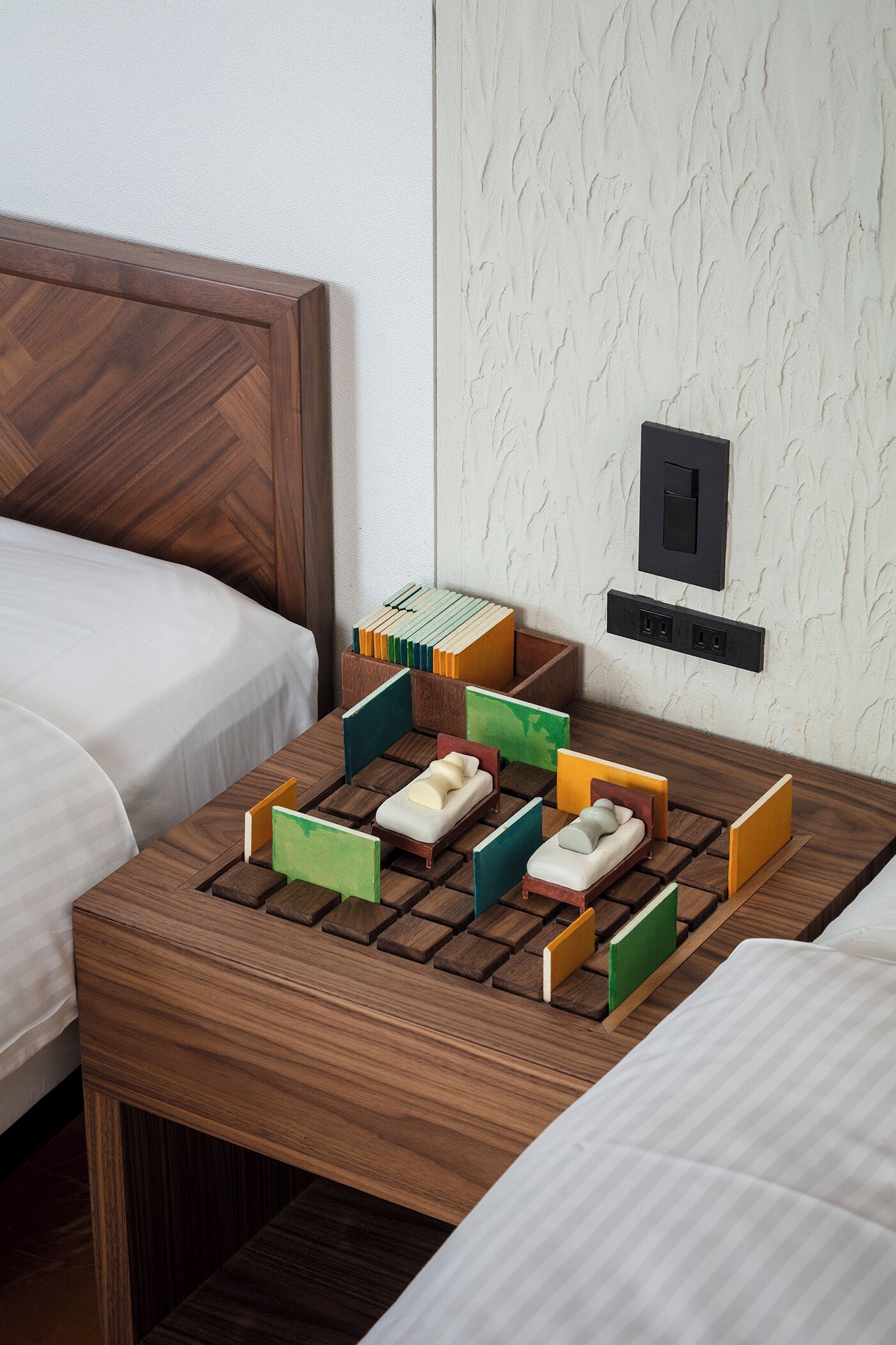A hotel room with two beds, a wooden nightstand with a board game, and a textured wall.