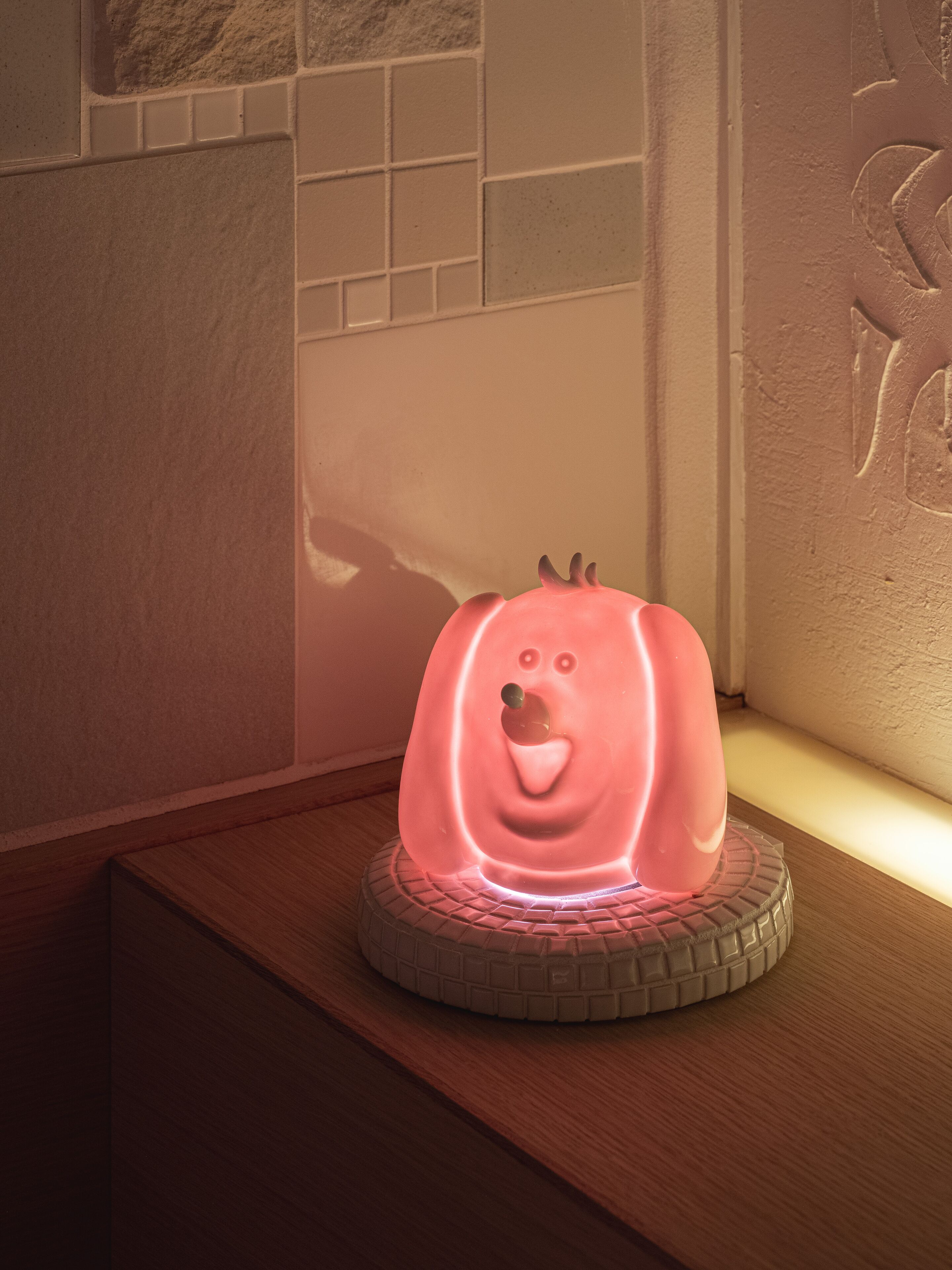 A pink, illuminated object resembling a pig on a circular base, placed on a wooden surface against a wall with a tiled section and a textured wall.