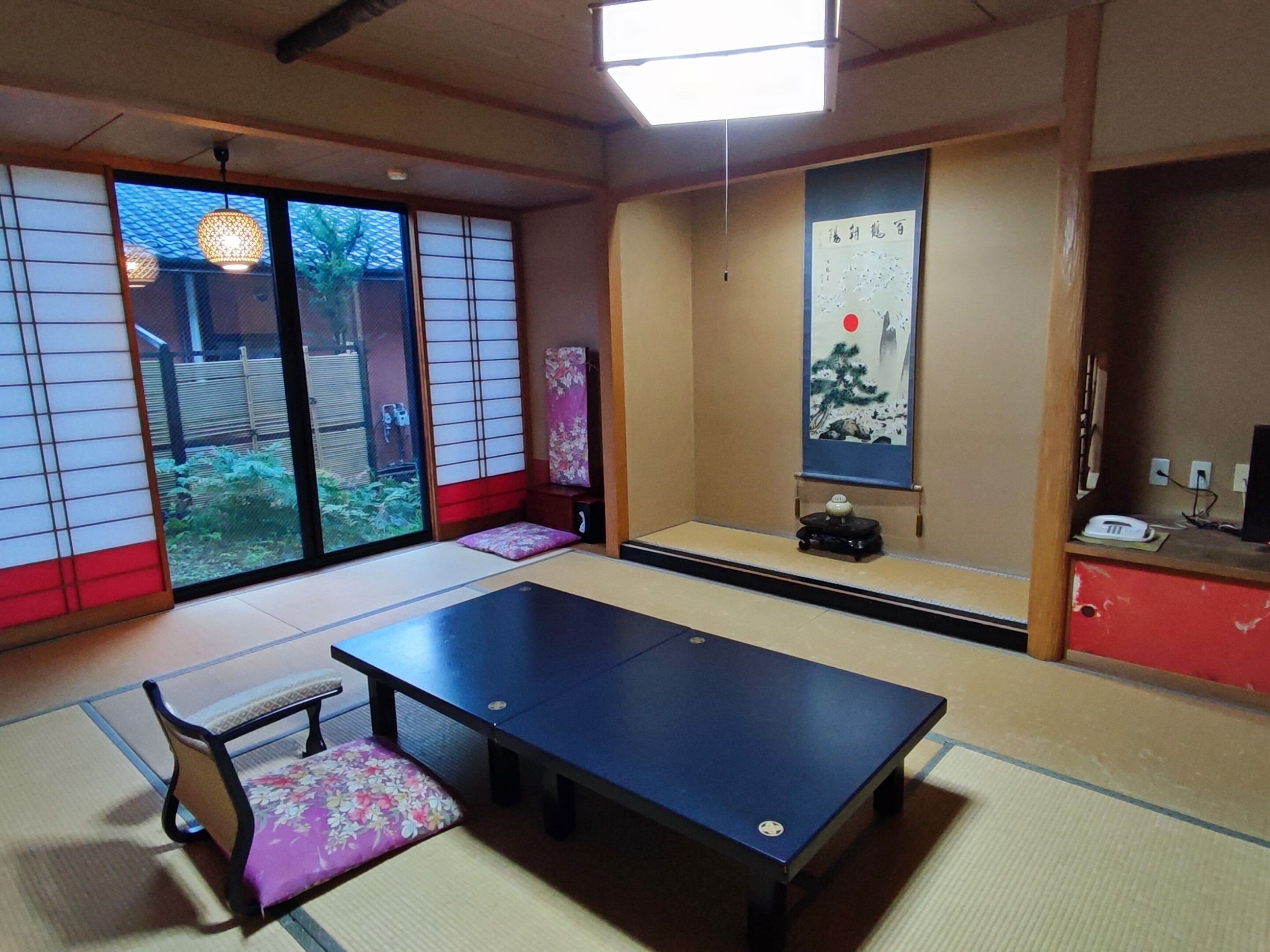 Una habitación con esteras de tatami, una mesa de tatami, un banco de madera bajo, una puerta corredera hacia un jardín y una pintura tradicional japonesa en la pared.