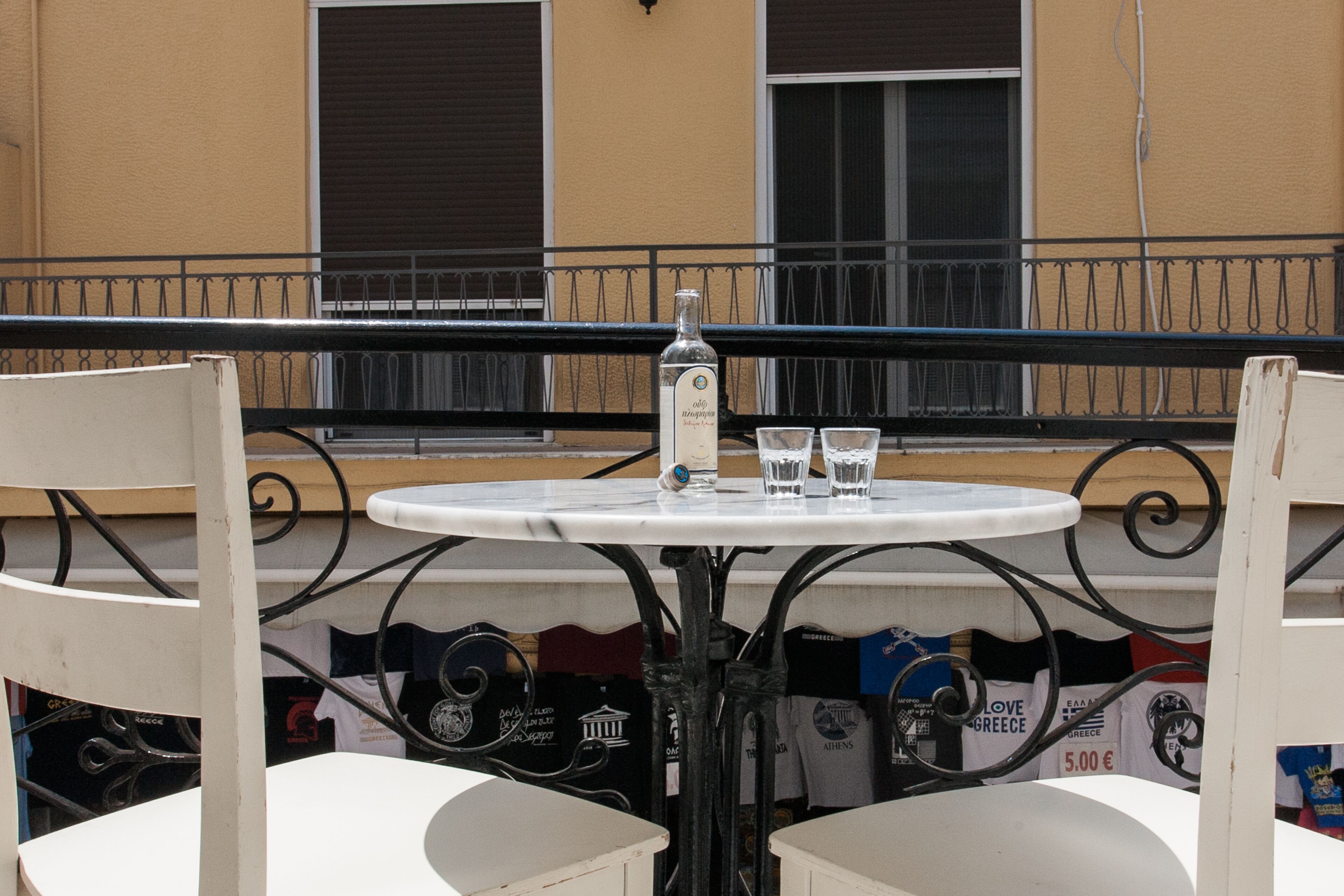 Una mesa redonda con una botella y dos vasos, rodeada de sillas blancas, frente a una fachada de edificio con carteles y una barandilla de metal negro.