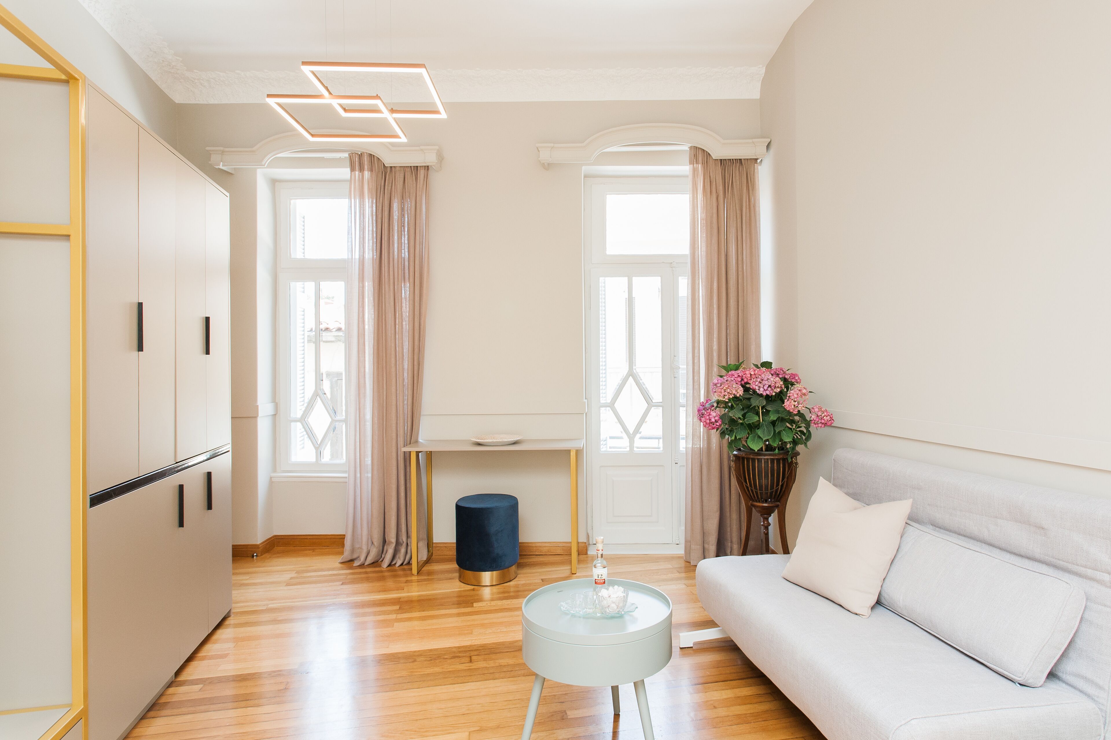 Una sala de estar moderna con un sofá, una mesita redonda y un reposapiés azul. Hay dos ventanas con cortinas, un piso de madera y una pared blanca con una lámpara geométrica.