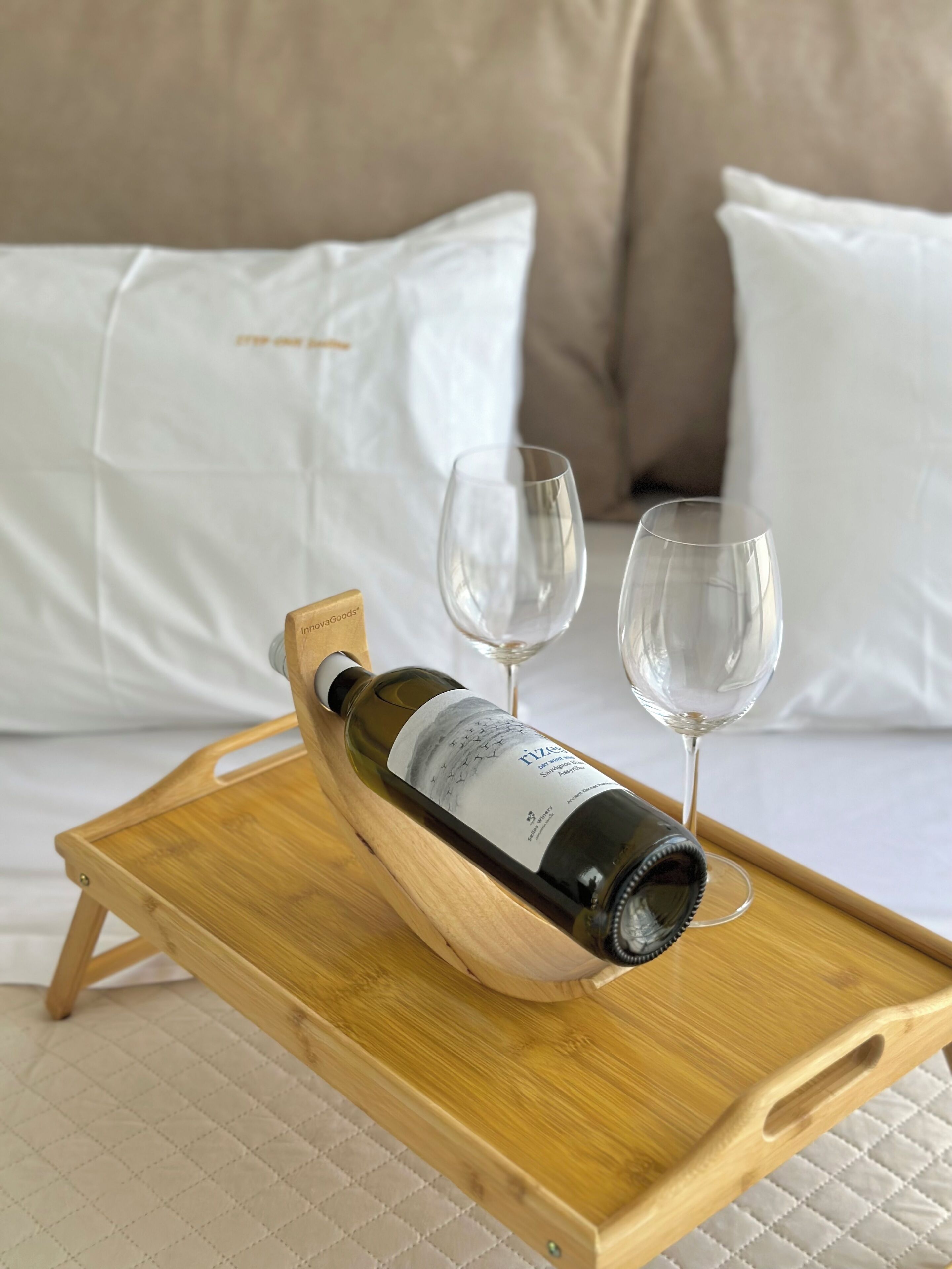 Una botella de vino sobre una bandeja de madera con dos copas vacías en una cama con almohadillas blancas.
