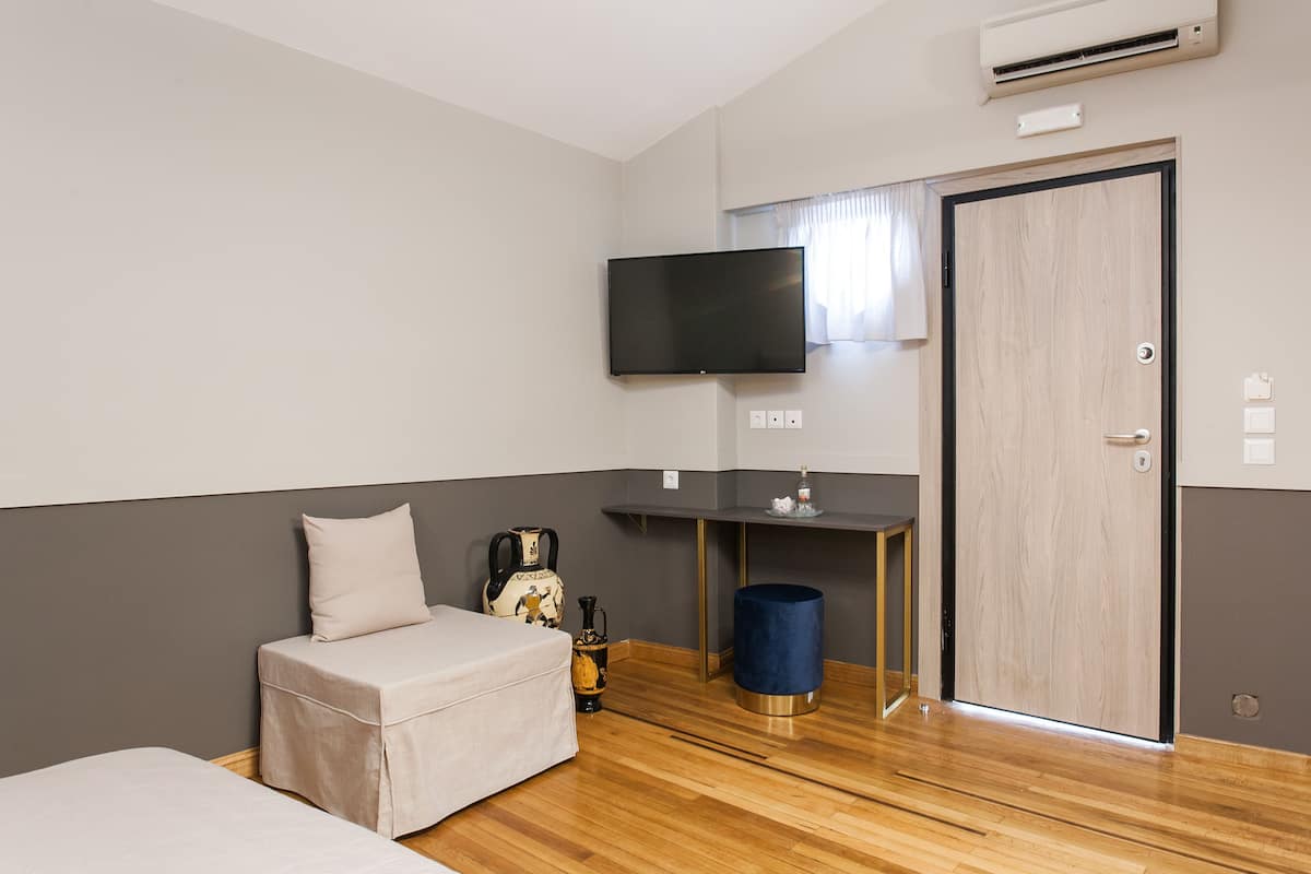 A room with a bed, a small table with a kettle, a blue trash bin, a wooden door, and a mounted TV.