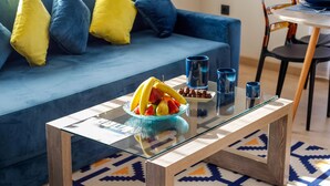 A modern living room with a glass coffee table, a blue sofa with yellow cushions, and a dining area with chairs and a table.