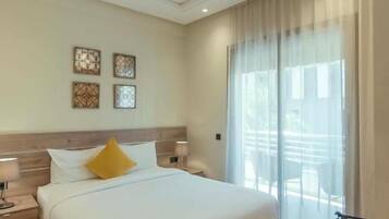 A modern hotel room with a large bed, wooden headboard, and a view of the outdoors through sheer curtains.