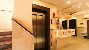 A hotel reception area with a marble staircase, an elevator, a reception desk with a red sign, and a ceiling fan. - GD House (Gurugram)