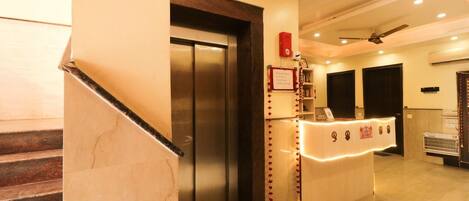 A hotel reception area with a marble staircase, an elevator, a reception desk with a red sign, and a ceiling fan.
