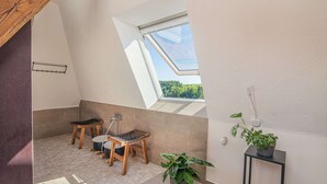 A small, sunlit room with a skylight, a wooden stool, a potted plant, and a black side table.