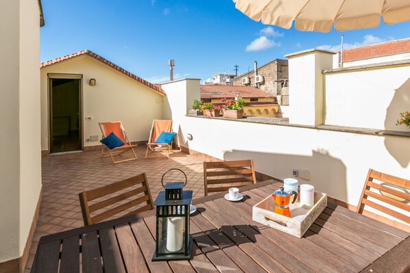 A rooftop terrace with wooden furniture, a table with a coffee pot, and potted plants.